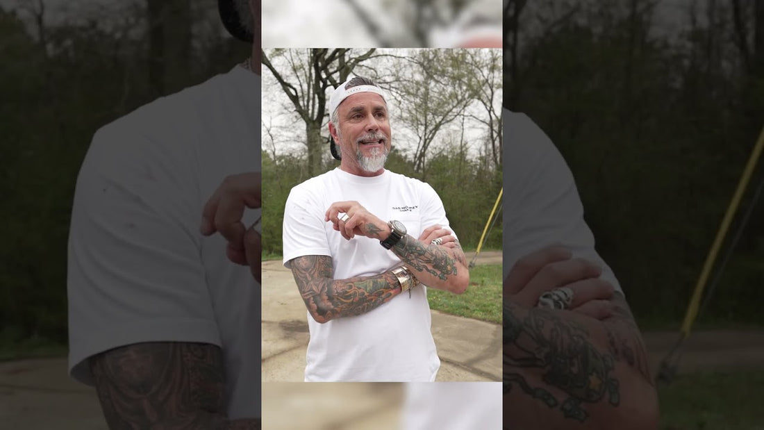 Tattooed man in a white Gas Monkey Garage T shirt and backward cap standing outdoors with arms crossed