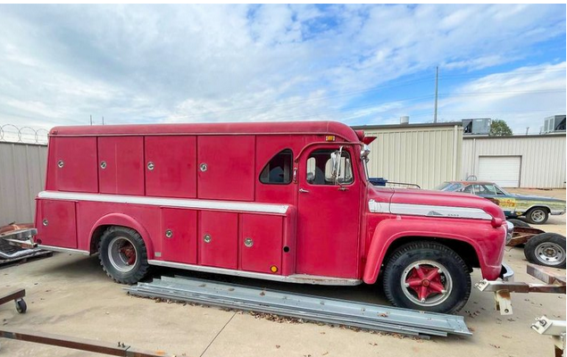 Side view of 1959 Ford F-700 fire truck showcasing its red body and side compartments with an industrial building in the background