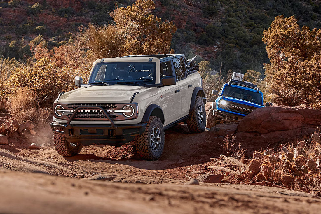 Two Ford Broncos on a rocky off-road trail with wooded hills in the background